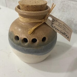May include: A small, round ceramic garlic keeper with a cork stopper. The keeper is tan, blue, and white with several holes for ventilation. A tag is tied around the neck with raffia. The text on the tag is blurry.