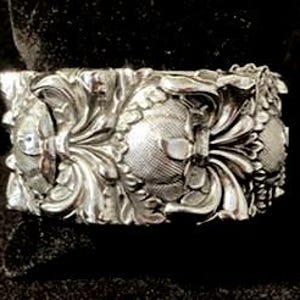 May include: A silver-toned cuff bracelet with intricate floral and foliate detailing. The design features raised patterns of leaves, flowers, and textured circular elements. The bracelet has a rectangular shape and appears to be made of metal.