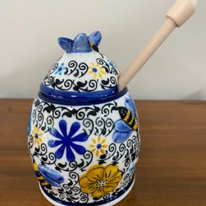 May include: A ceramic honey pot with a wooden dipper. The pot is white with blue, yellow, and black floral and bee designs. The lid has a blue flower-shaped handle. The dipper is inserted into the pot.