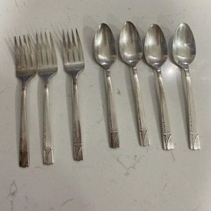 May include: A set of vintage silverware, including three forks and three spoons. The silverware is silver-colored with a decorative pattern on the handles. The spoons have oval bowls, and the forks have four tines.