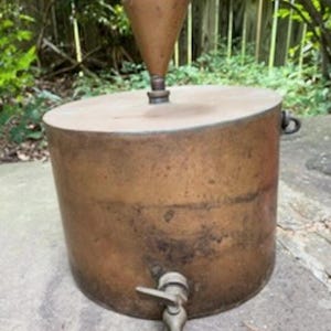 May include: A cylindrical copper vessel featuring a flat top lid with a central conical funnel attachment. The body of the container includes a metal spigot near the base and a small side handle. The exterior surface shows a weathered patina.