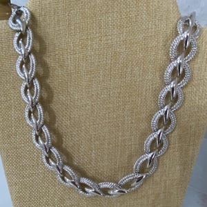 May include: A silver-toned chain necklace featuring a series of interlocking oval links. Each link displays a textured, cross-hatched pattern on the outer edges, contrasted by smooth, polished silver-colored sections at the connection points.