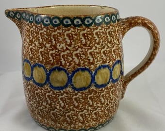 May include: A ceramic pitcher with a brown and white speckled pattern. The pitcher has a handle and spout, with blue and yellow circular designs around the body and green accents at the rim.
