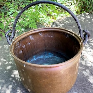 May include: A copper cauldron with a dark, wrought iron handle. The pot has a wide opening and a rounded base. The interior shows signs of use with a blue-green patina. The cauldron is outdoors, with greenery in the background.