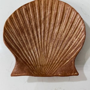 May include: A ceramic dish shaped like a scallop shell with a textured, ribbed surface. The dish features a warm, earthy brown color with darker brown speckles throughout, mimicking the natural appearance of a seashell in a functional form.