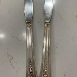 May include: Two silver butter knives with ornate floral designs on the handles. The blades are slightly curved and the handles have a detailed pattern. The knives are displayed on a light-colored surface.
