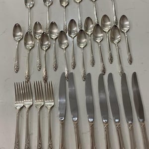 May include: A collection of silver-colored flatware, including spoons, forks, and knives, arranged on a light-colored surface. The spoons have a rounded bowl and ornate handle detailing. The forks and knives also feature decorative handles, suggesting a formal dining set.