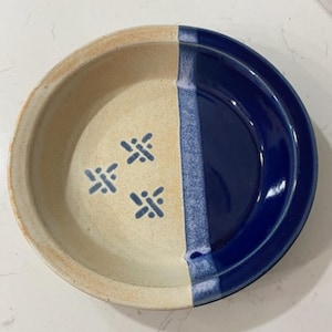 May include: A round ceramic bowl featuring a split design with one half in a matte beige finish decorated with three small blue cross-shaped patterns and the other half in a glossy deep blue finish, separated by a textured blue vertical band.