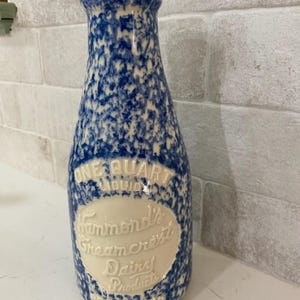 May include: A blue and white ceramic milk bottle with the text "ONE QUART LIQUID" and "Hammond's Creamcrest Dairy Products REGISTERED". The bottle has a speckled design and a narrow neck, reminiscent of vintage dairy containers.