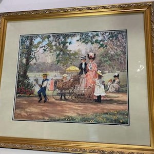 May include: A framed print depicting a park scene with figures in period clothing. The artwork features a gold ornate frame, a light-colored mat, and a detailed illustration of people with carriages and children in a park setting. The colors are soft and muted.