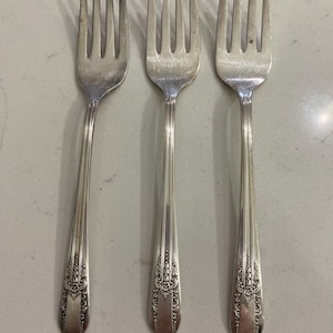 May include: Three silver forks with ornate floral detailing on the handles. The forks have tines and a polished finish, suggesting they are part of a formal dining set. The design is classic and elegant.