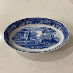 May include: Oval serving platter with a blue and white landscape design. The rim features a floral pattern. The center of the platter depicts a pastoral scene with trees, buildings, and figures. A classic piece for serving food.