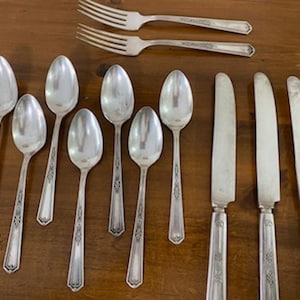 May include: A collection of vintage silverware, including spoons, forks, and knives, arranged on a wooden surface. The silverware has a silver finish with decorative detailing on the handles. The spoons are oval-shaped, and the forks have four tines.