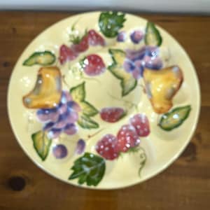 May include: A yellow ceramic serving plate with a fruit design. The plate features painted grapes, strawberries, and leaves. Two yellow, curved handles are on opposite sides of the plate.