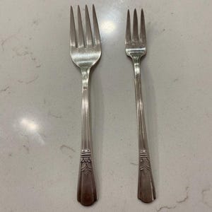 May include: Two silver forks with four tines each. The forks have long, slender handles with a decorative floral design at the base. The forks are placed side by side on a light-colored surface.