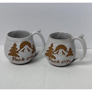 May include: Two round, light gray ceramic mugs with a glossy finish. Each mug features a brown graphic of a mountain range, two pine trees, and a sun, with the text Park City printed below the design. Each mug has a curved handle with a small tab.