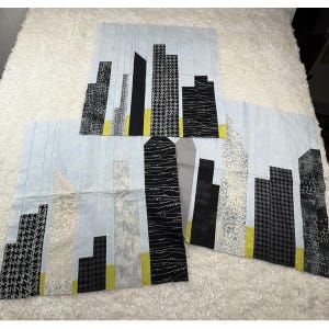 May include: Three rectangular fabric panels featuring a pieced city skyline design. The buildings are constructed from various black, white, and gray patterned fabrics, including houndstooth, dots, and lines, set against a light blue sky background.