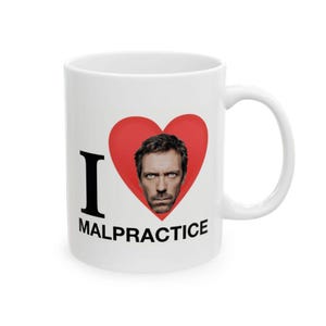 Puede incluir: Taza de cerámica blanca con una "I" negra y la palabra "MALPRACTICE" debajo de un corazón rojo que contiene un retrato de un hombre con ojos azules. La taza tiene un asa curva.