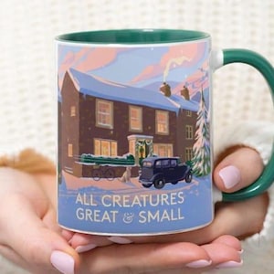 May include: A ceramic mug with a green handle and rim, featuring a winter scene with a house, car, and text "ALL CREATURES GREAT & SMALL". The mug is held by a person wearing a white sweater.