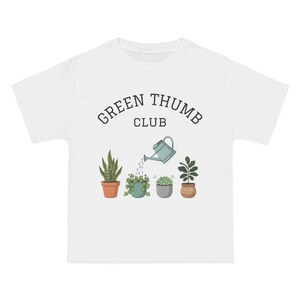 May include: White t-shirt with a graphic of four potted plants and a watering can. The text "GREEN THUMB CLUB" is arched above the plants. The plants are in terracotta, gray, and woven pots.