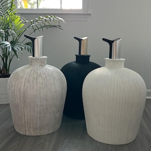 May include: Three decorative ceramic soap dispensers in neutral tones. The dispensers have a textured, ribbed design and silver or gold-toned pump tops. One is light gray, one is black, and one is off-white. The dispensers are on a gray wood-look floor.
