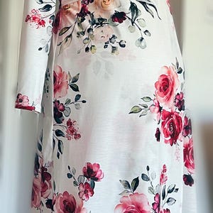 May include: White long-sleeved dress with a vibrant floral pattern of large pink and red roses, green leaves, and smaller floral accents. The dress has a V-neck and a fitted waist, creating a feminine silhouette.