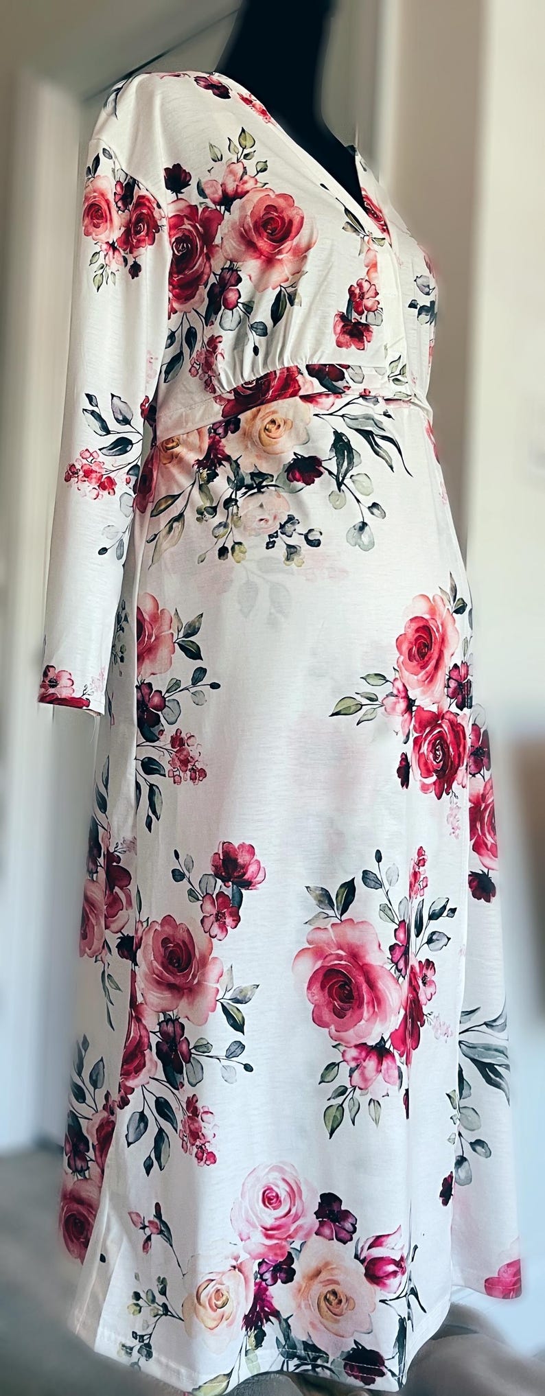 May include: White long-sleeved dress with a vibrant floral pattern of large pink and red roses, green leaves, and smaller floral accents. The dress has a V-neck and a fitted waist, creating a feminine silhouette.