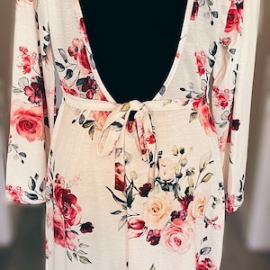 May include: A white dress with a floral pattern of red and pink roses and green leaves. The dress has long sleeves, a low back, and a tie waist. It is displayed on a black mannequin.
