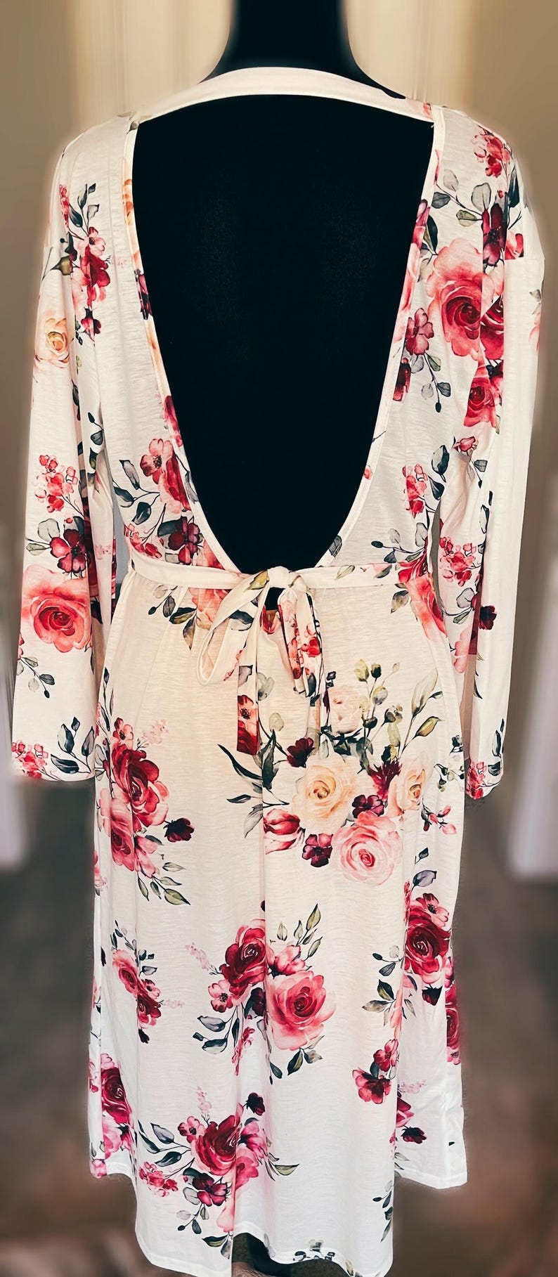 May include: A white dress with a floral pattern of red and pink roses and green leaves. The dress has long sleeves, a low back, and a tie waist. It is displayed on a black mannequin.