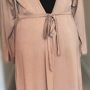 May include: A long, pale brown robe with long sleeves and a deep V-neck back. The robe is tied at the waist with a matching fabric belt. The fabric appears soft and flowing, suitable for lounging or as a cover-up.