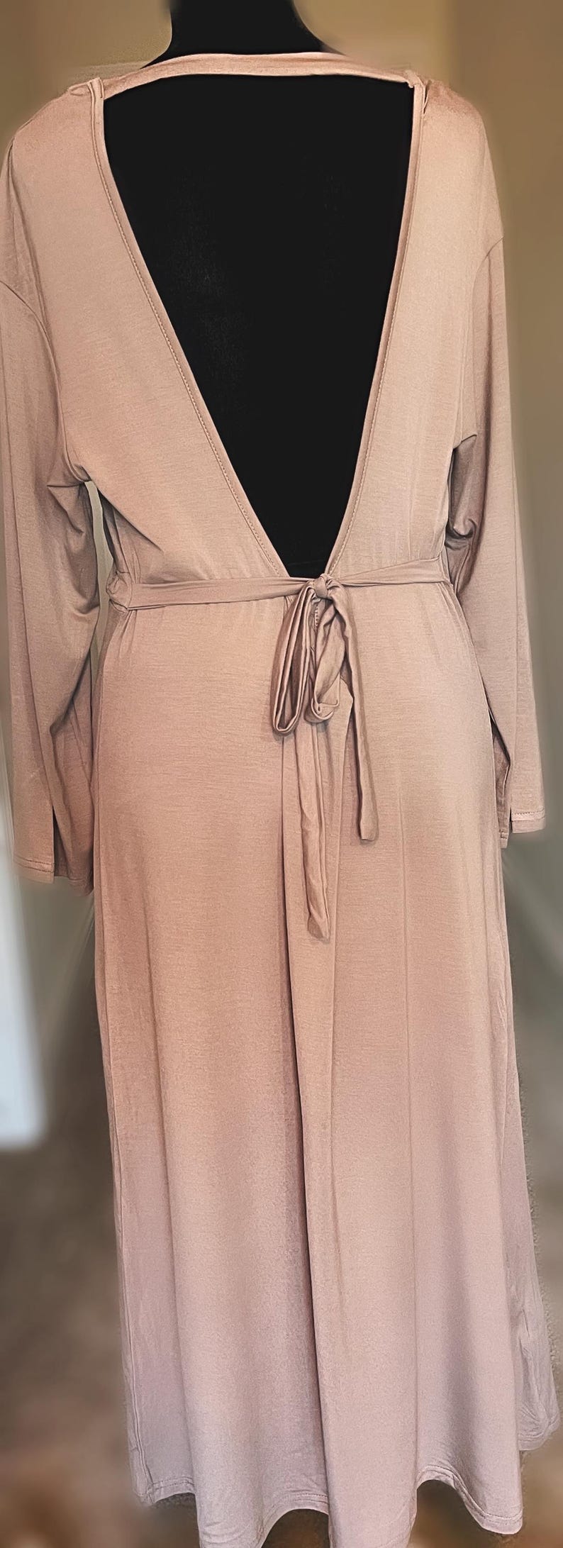May include: A long, pale brown robe with long sleeves and a deep V-neck back. The robe is tied at the waist with a matching fabric belt. The fabric appears soft and flowing, suitable for lounging or as a cover-up.