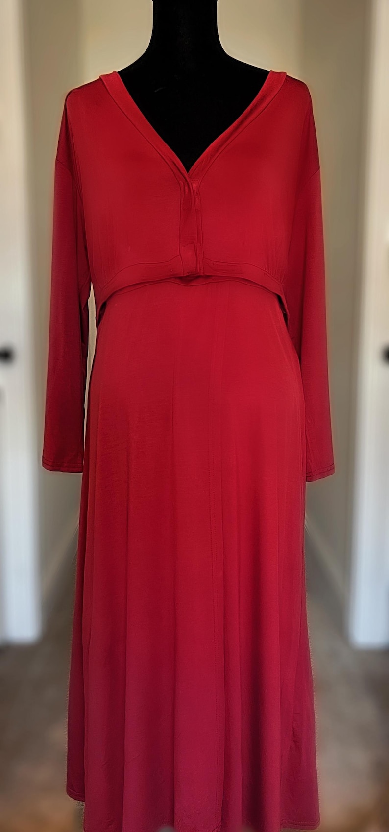 May include: A long, red dress with long sleeves and a V-neckline. The dress has a button closure at the bust and a flowing skirt. The dress is displayed on a black mannequin.