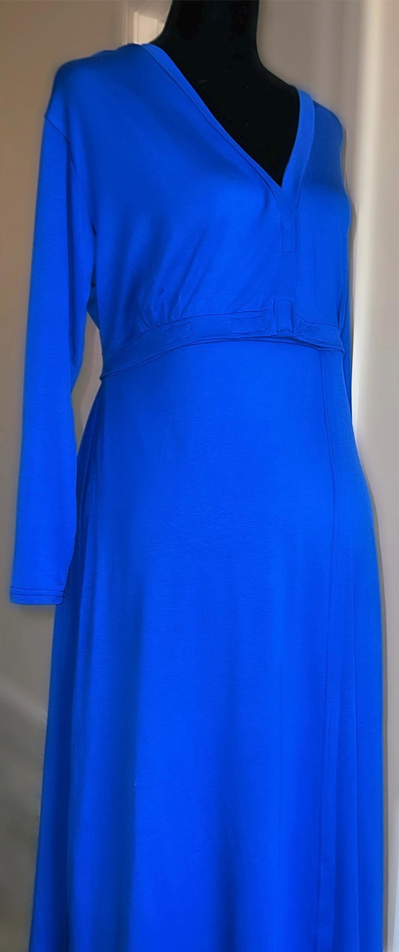May include: A long, royal blue dress with long sleeves. The dress features a V-neckline and a fitted waist with a horizontal band. The fabric drapes smoothly, creating a flowing silhouette. The dress is displayed on a black mannequin.