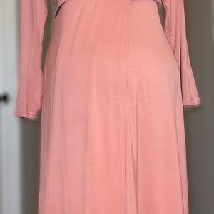 May include: A long-sleeved, dusky pink dress with a V-neck and button fastening. The dress has a fitted waist and a flowing skirt, creating a simple yet elegant silhouette. The dress is displayed on a black mannequin.