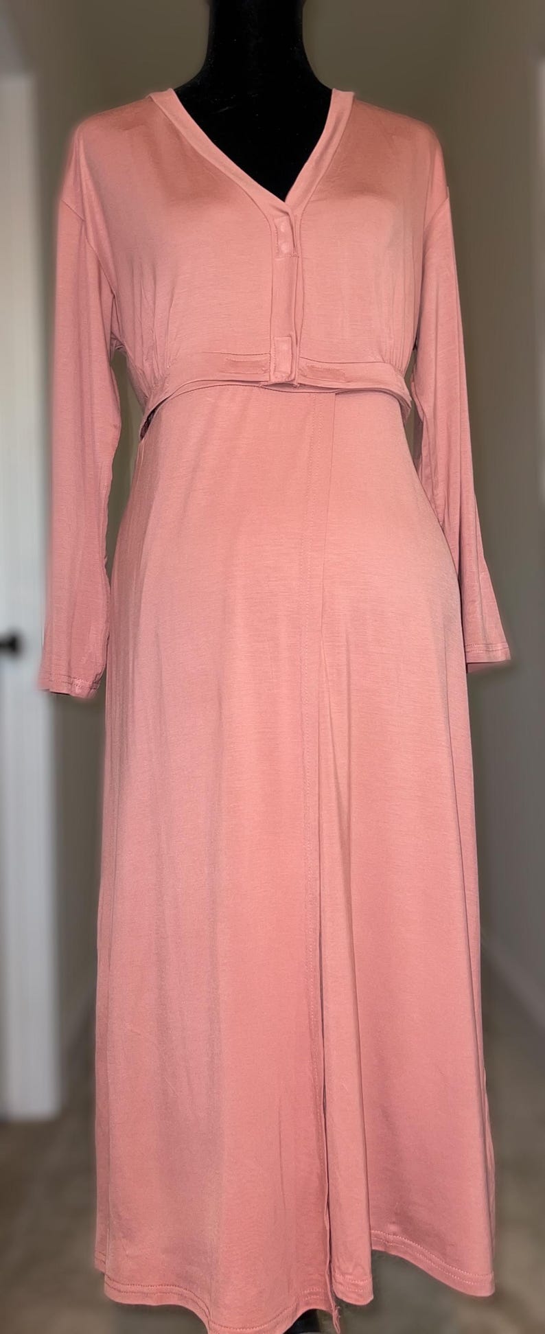 May include: A long-sleeved, dusky pink dress with a V-neck and button fastening. The dress has a fitted waist and a flowing skirt, creating a simple yet elegant silhouette. The dress is displayed on a black mannequin.