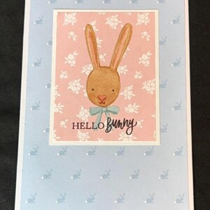 May include: A greeting card featuring a watercolor bunny illustration. The bunny has long ears, a blue bow, and the words "Hello Bunny" written below. The card has a light blue background with bunny silhouettes.