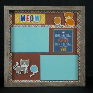 May include: A framed cat-themed shadow box with a gray wooden frame. The box features two blue photo mats, brown backing, and decorative cat stickers. Text includes "MEOW" and "First they steal your heart then they steal your bed."