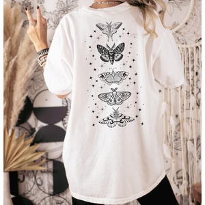 May include: White t-shirt featuring a black graphic of five moths in a vertical arrangement, each with intricate wing patterns. Small star shapes are scattered around the moths. The shirt is a casual style, suitable for everyday wear.