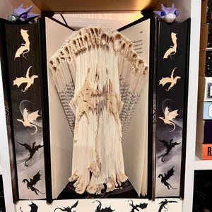 May include: A unique book art piece featuring a castle design carved from the pages of a book. The book is flanked by two black bookends with dragon silhouettes. Two purple figurines sit on top of the bookends. The shelf also has dragon silhouettes.