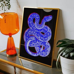 Peut inclure: Une impression d'art encadrée représentant une illustration de serpent bleu et blanc sur fond noir. L'œuvre est dans un cadre doré et est exposée sur une table bordée d'or. Une lampe orange et une plante en pot sont également visibles.