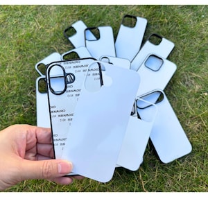 Peut inclure: Plusieurs coques de téléphone blanches avec des bords noirs, certaines avec un film protecteur. Les coques sont disposées sur de l'herbe verte. On peut lire "CROWN #512" sur certaines coques.