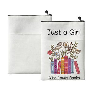 Puede incluir: Dos bolsas de lona blancas con cremalleras negras y bolsillos delanteros. Una presenta el texto "Just a Girl Who Loves Books" con un diseño floral y de libros. La otra bolsa es lisa.