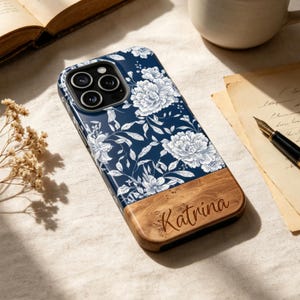 Puede incluir: Una funda para smartphone con fondo azul oscuro, un patrón floral blanco en la parte superior y una textura de veta de madera clara en la parte inferior, con el nombre Katrina grabado en cursiva en la sección de madera.