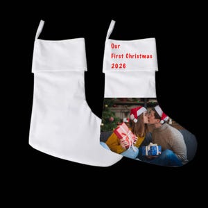 Puede incluir: Dos calcetines navideños blancos con un lazo para colgar. Uno es blanco liso, mientras que el otro presenta el texto rojo Our First Christmas 2026 y una fotografía impresa de dos personas con gorros de Papá Noel intercambiando regalos.