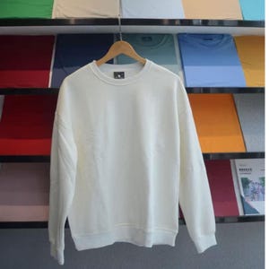 May include: A cream-colored sweatshirt with a crew neck and long sleeves hangs on a wooden hanger. The sweatshirt is displayed against a backdrop of folded shirts in various colors, including blue, green, and orange.
