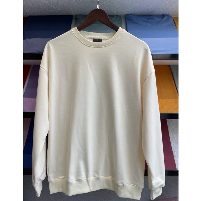 May include: Cream-colored sweatshirt with a crew neck and long sleeves. The garment is hanging on a wooden hanger. The background features folded shirts in various colors.
