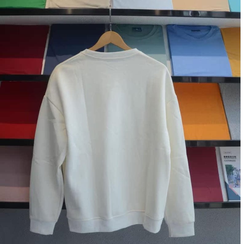 May include: Cream-colored sweatshirt with a crew neck and long sleeves, hanging on a wooden hanger. The sweatshirt is displayed against a backdrop of folded t-shirts in various colors, including blue, green, and orange.
