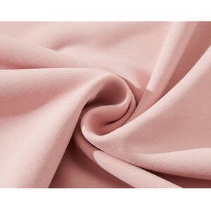 May include: Close-up of a soft, blush pink fabric. The material is tightly woven with a subtle texture, and it is draped in gentle folds, creating shadows and highlights. The fabric appears smooth and luxurious.