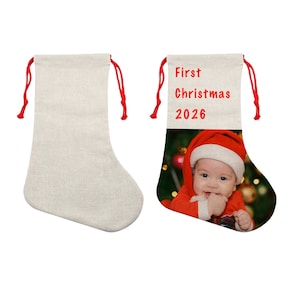 Puede incluir: Dos calcetines de tela beige con cordones rojos. Uno es liso, mientras que el otro presenta el texto rojo impreso First Christmas 2026 sobre una fotografía de un bebé con un gorro de Papá Noel y ropa de color rojo.