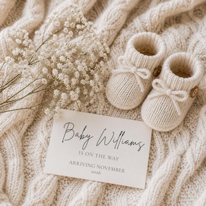 May include: A pair of cream-colored knit baby booties with small brown buttons and bows, accompanied by a sprig of white baby's breath flowers and a card that reads Baby Williams IS ON THE WAY ARRIVING NOVEMBER 2026.